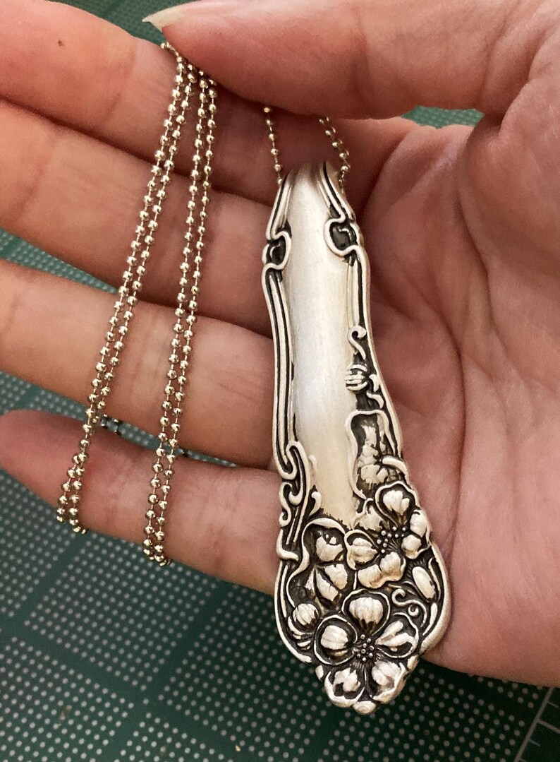 May include: A silver necklace with a pendant made from a vintage spoon. The spoon has a floral design and is attached to a silver ball chain.
