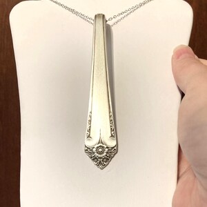 May include: Silverware pendant necklace. The pendant is a repurposed spoon with a floral design at the base. The necklace is displayed on a white stand. The chain is a delicate silver color.