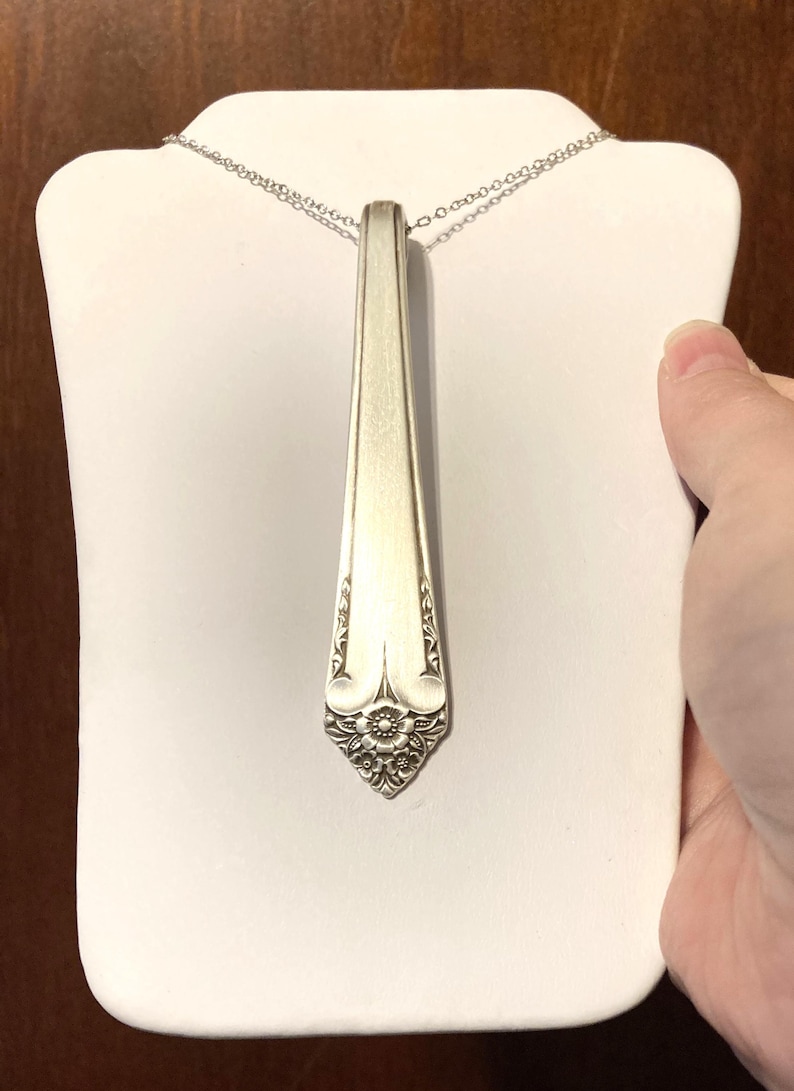 May include: Silverware pendant necklace. The pendant is a repurposed spoon with a floral design at the base. The necklace is displayed on a white stand. The chain is a delicate silver color.