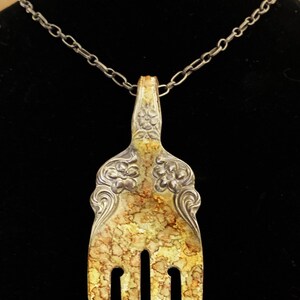 May include: A silver fork necklace with a decorative design and a yellow patina. The fork is attached to a silver chain.