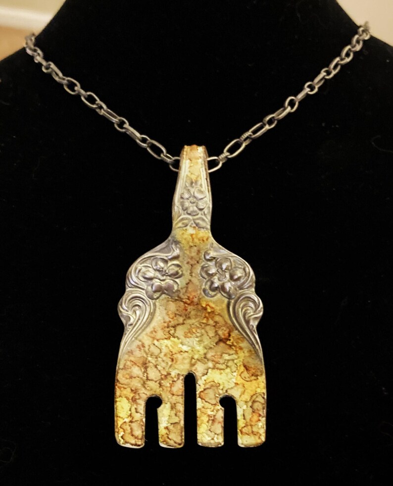 May include: A silver fork necklace with a decorative design and a yellow patina. The fork is attached to a silver chain.