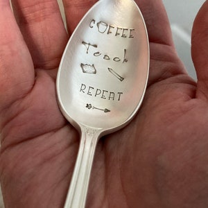 May include: A silver spoon with the words "Coffee, Teach, Repeat" engraved on it. There are also small drawings of a coffee cup, a book, a pencil, and an arrow.