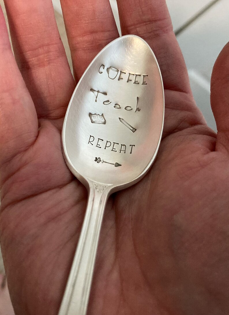 May include: A silver spoon with the words "Coffee, Teach, Repeat" engraved on it. There are also small drawings of a coffee cup, a book, a pencil, and an arrow.