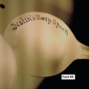 May include: A gold-colored soup spoon with the words "Justin's Soup Spoon" engraved on the front. The spoon is shown against a light brown background. Font #4.