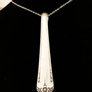 May include: A silver-colored pendant necklace featuring a repurposed spoon head. The spoon's handle is attached to a delicate chain. The spoon head has an ornate floral and scroll design at the base, adding a vintage touch to the jewelry.