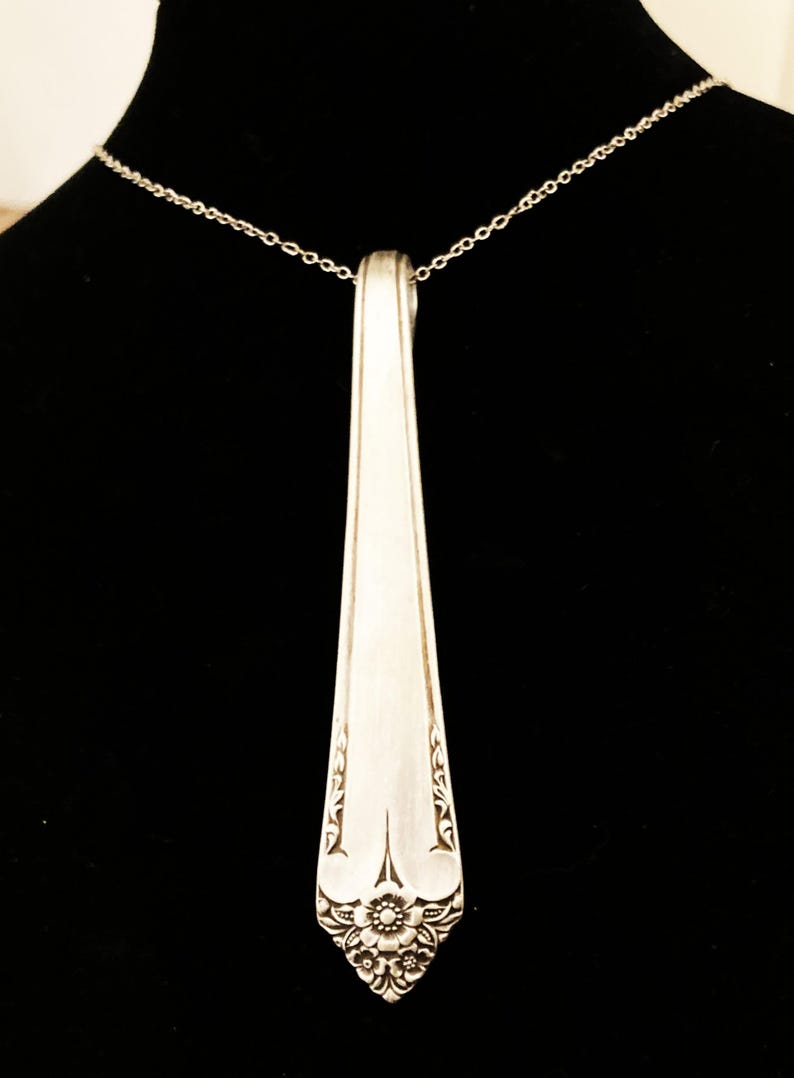 May include: A silver-colored pendant necklace featuring a repurposed spoon head. The spoon's handle is attached to a delicate chain. The spoon head has an ornate floral and scroll design at the base, adding a vintage touch to the jewelry.