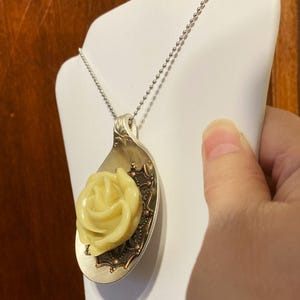 May include: A silver spoon necklace with a yellow rose charm. The spoon has a decorative design around the edge.