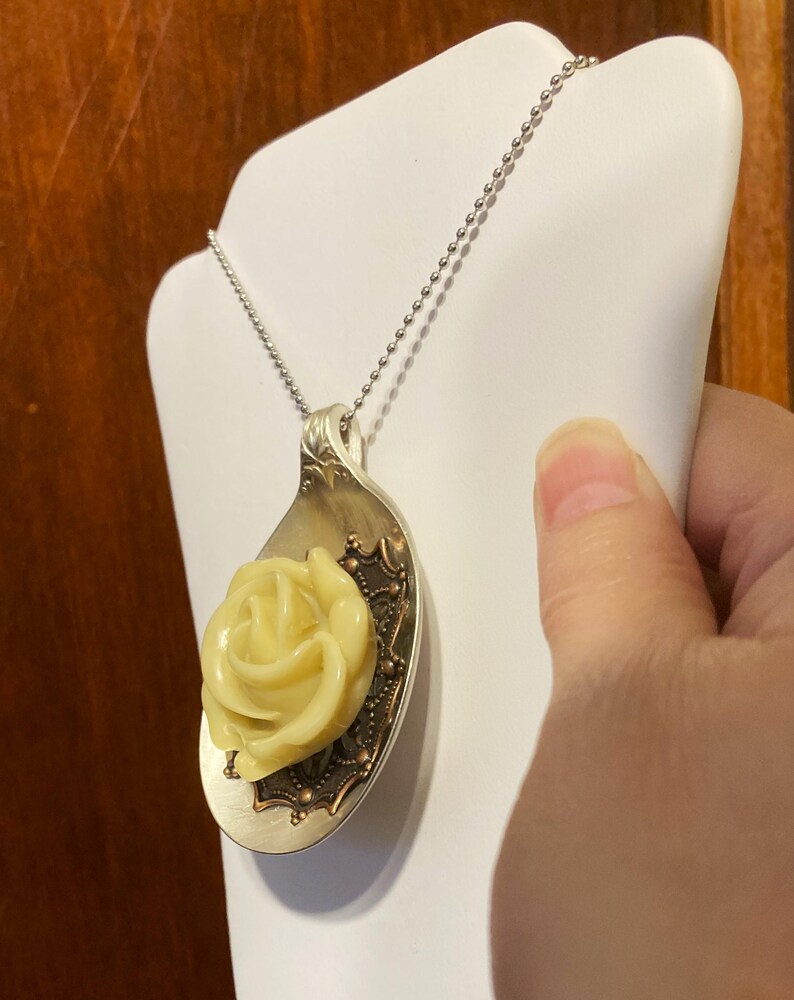 May include: A silver spoon necklace with a yellow rose charm. The spoon has a decorative design around the edge.