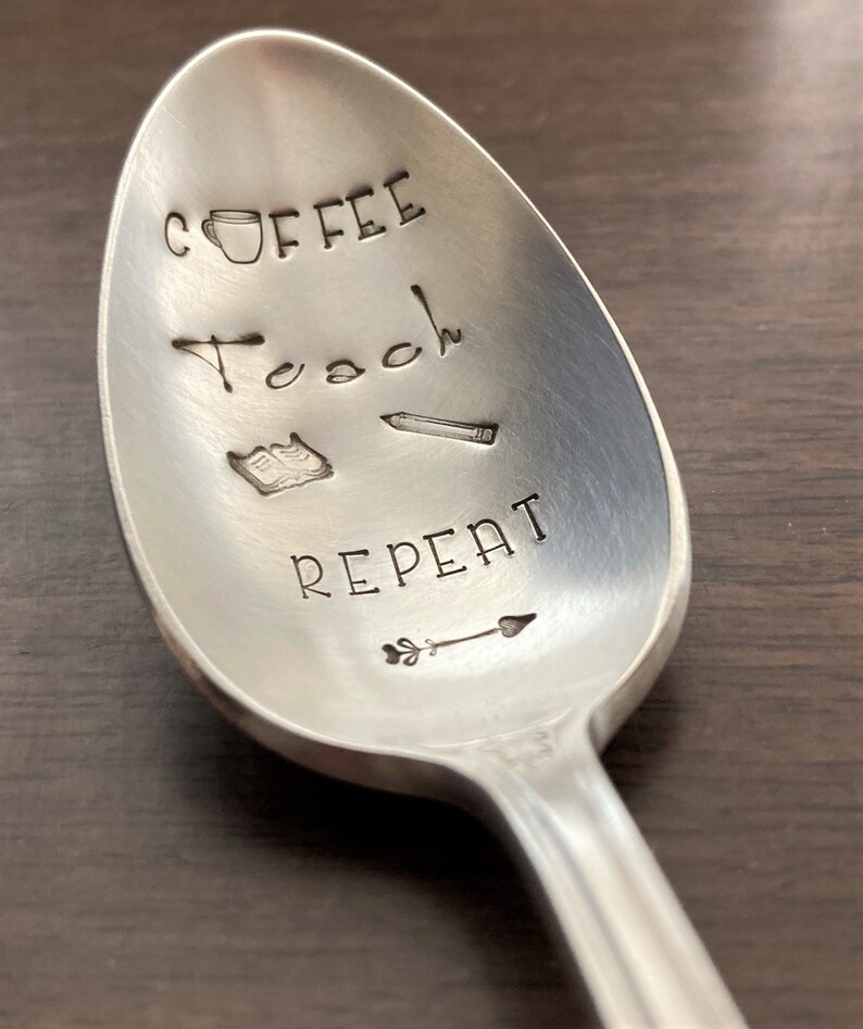 May include: A silver spoon with the words "Coffee Teach Repeat" engraved on it. The spoon also has a small coffee cup, a book, a pencil, and an arrow engraved on it.