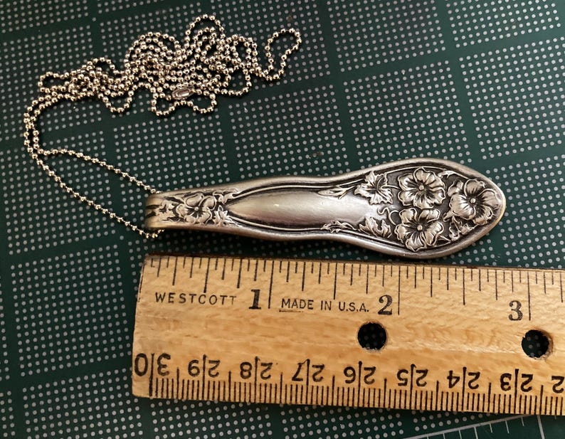 May include: A silver necklace with a floral design. The pendant is a vintage silverware spoon with a floral pattern. The necklace is 2 inches long.