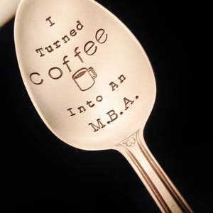 Pu&ograve; includere: Cucchiaio d'argento con l'incisione "I turned coffee into an M.B.A." incisa nella coppetta. Il cucchiaio ha un manico decorativo e un disegno di tazza di caff&egrave;. Lo sfondo &egrave; nero.