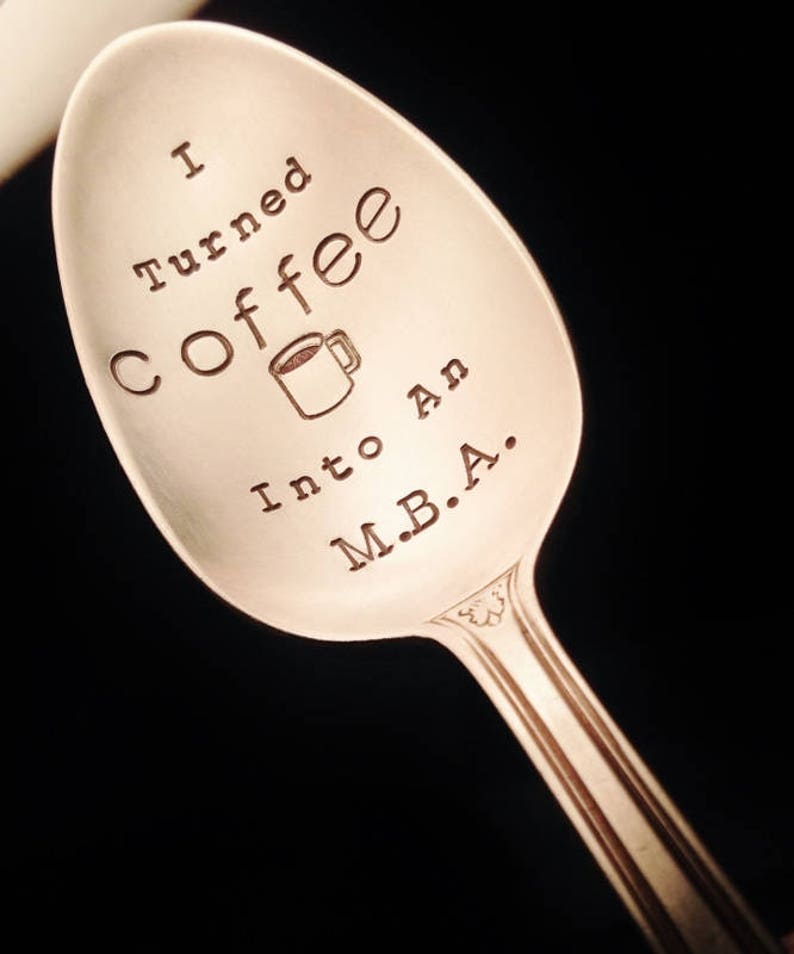 Pu&ograve; includere: Cucchiaio d'argento con l'incisione "I turned coffee into an M.B.A." incisa nella coppetta. Il cucchiaio ha un manico decorativo e un disegno di tazza di caff&egrave;. Lo sfondo &egrave; nero.
