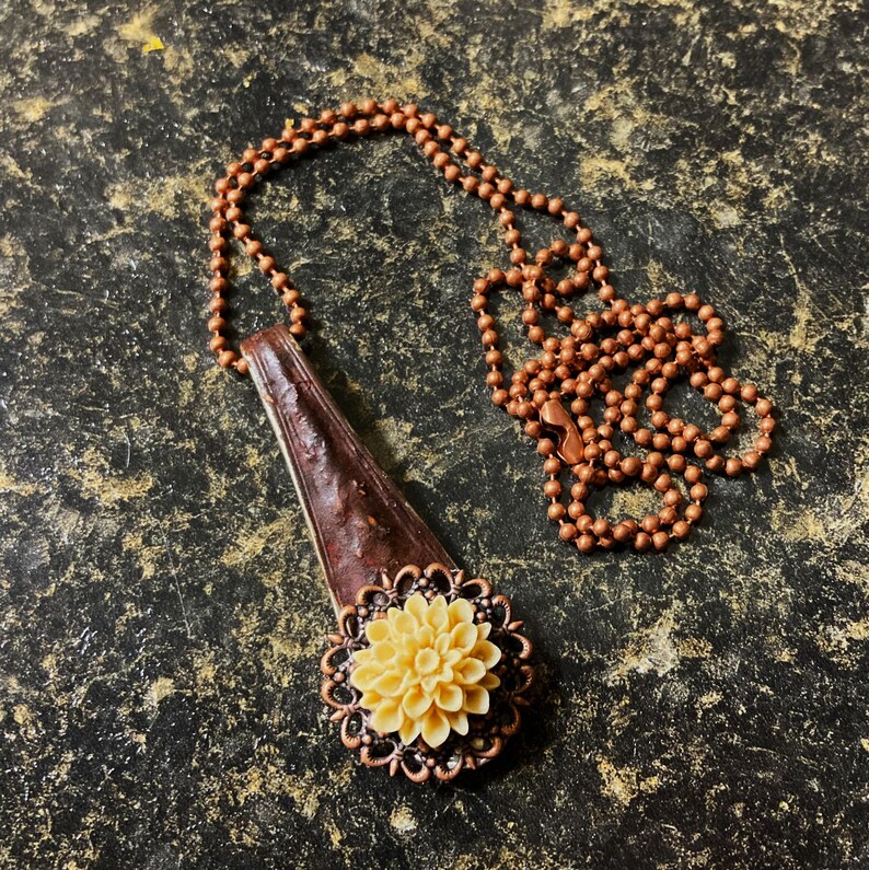 May include: A copper necklace with a brown leather pendant featuring a cream-colored flower with a copper frame.