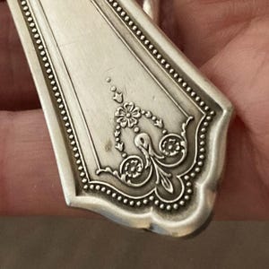 May include: Close-up of the ornate, silver-plated handle of a vintage spoon. The handle features a detailed floral design with a beaded border.