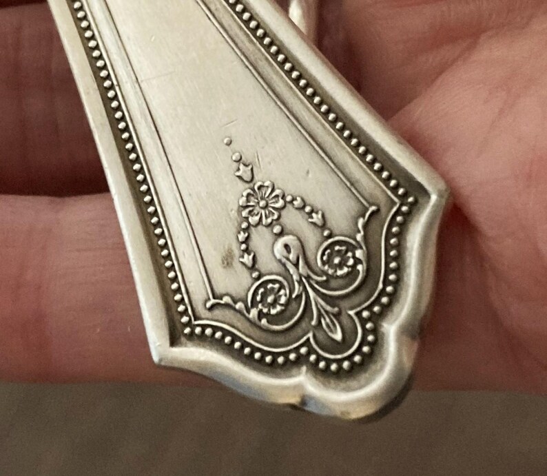 May include: Close-up of the ornate, silver-plated handle of a vintage spoon. The handle features a detailed floral design with a beaded border.