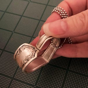 May include: A silver spoon bent into a heart shape with a chain attached. The spoon has an ornate design on the handle.