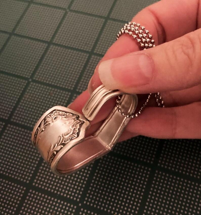 May include: A silver spoon bent into a heart shape with a chain attached. The spoon has an ornate design on the handle.