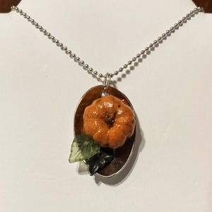 May include: A silver chain necklace with a pendant featuring a small orange pumpkin with green leaves on a brown oval background.