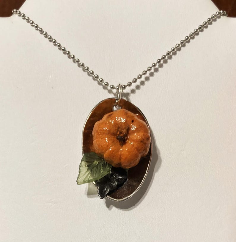 May include: A silver chain necklace with a pendant featuring a small orange pumpkin with green leaves on a brown oval background.