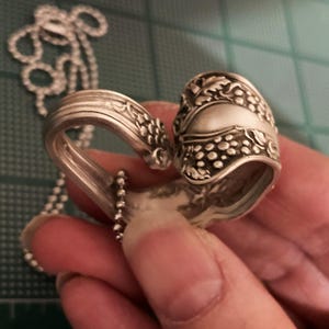 May include: A silver spoon necklace with a heart-shaped design. The spoon is decorated with a grapevine pattern and has a chain attached to it.