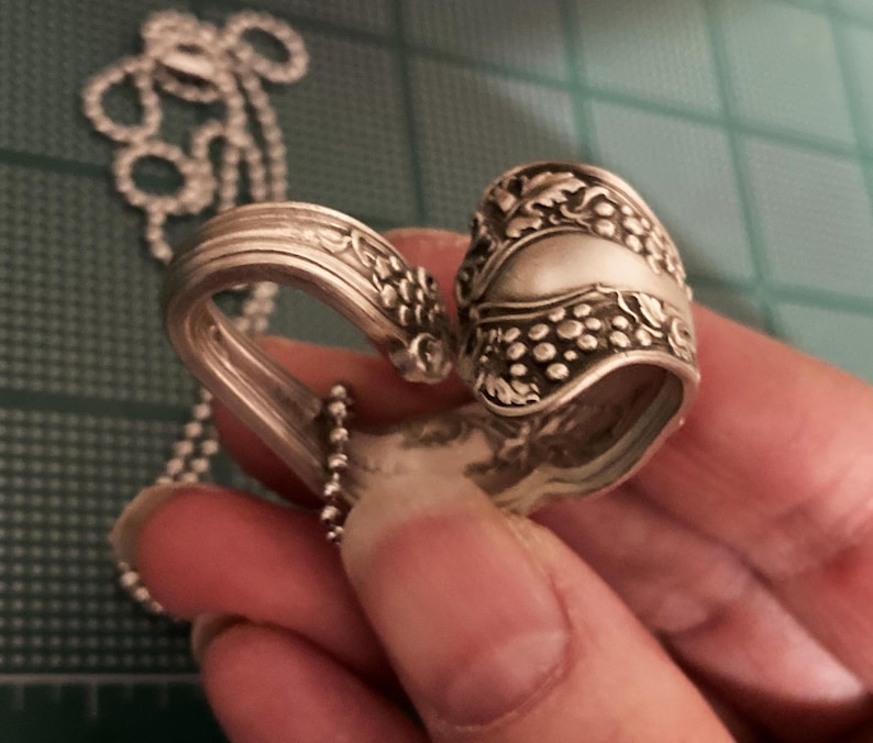 May include: A silver spoon necklace with a heart-shaped design. The spoon is decorated with a grapevine pattern and has a chain attached to it.