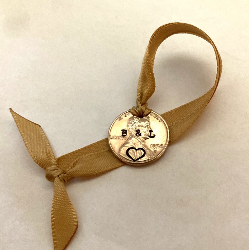 K&ouml;nnte beinhalten: Ein goldfarbener US-Penny mit dem Jahr 1974 eingraviert. Der Penny ist an einem goldenen Band mit einer Schleife gebunden. Die Initialen "B & L" sind zusammen mit einem Herzen in den Penny eingraviert.
