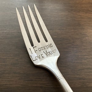 May include: A silver fork with the words "I ForKing Love You" engraved on the handle. The fork is on a wooden surface.