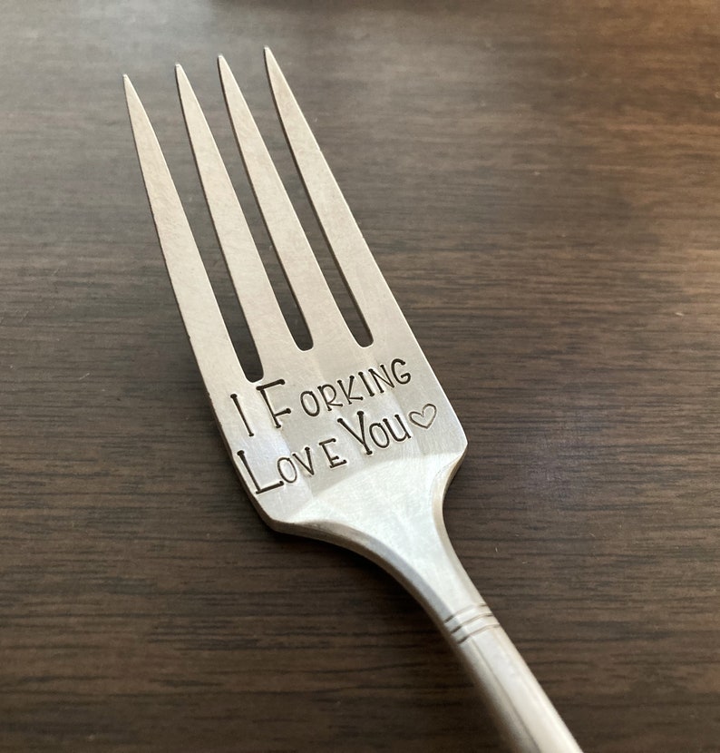 May include: A silver fork with the words "I ForKing Love You" engraved on the handle. The fork is on a wooden surface.