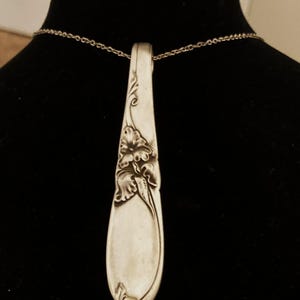 Puede incluir: Collar con colgante de cuchara plateada. El cuenco de la cuchara presenta un dise&ntilde;o floral. El colgante cuelga de una delicada cadena plateada. El mango de la cuchara es curvo y tiene detalles decorativos. El collar se muestra sobre un fondo negro.
