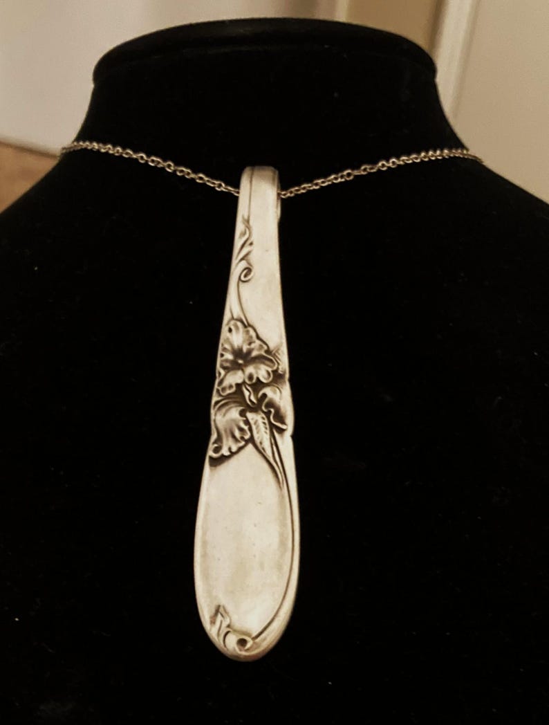 Puede incluir: Collar con colgante de cuchara plateada. El cuenco de la cuchara presenta un dise&ntilde;o floral. El colgante cuelga de una delicada cadena plateada. El mango de la cuchara es curvo y tiene detalles decorativos. El collar se muestra sobre un fondo negro.