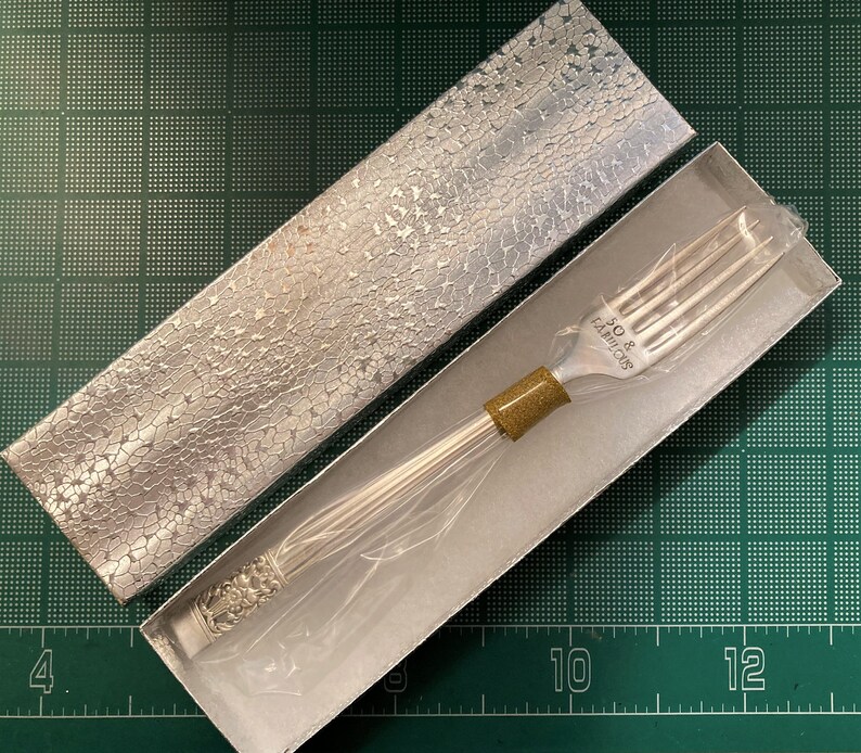 May include: A silver fork with a gold handle in a silver and white box. The fork has the words "30 & LA BELLE" engraved on the handle.