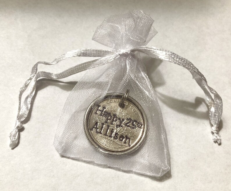 May include: A silver-colored coin pendant inside a sheer white organza gift bag. The coin has the inscription "Happy 25th Allison" and the year 1998. The bag is tied with a matching white ribbon.