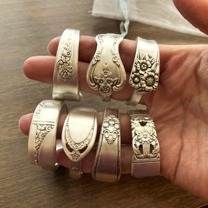 Puede incluir: Un conjunto de ocho pulseras de cuchara de plata con dise&ntilde;os florales. Las pulseras est&aacute;n dispuestas en una fila en una mano.