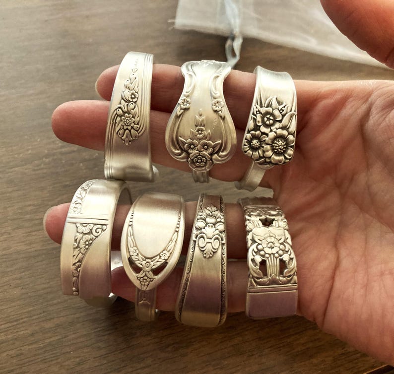 Puede incluir: Un conjunto de ocho pulseras de cuchara de plata con dise&ntilde;os florales. Las pulseras est&aacute;n dispuestas en una fila en una mano.