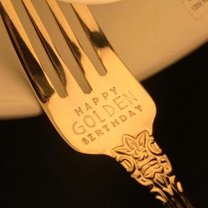 May include: Close-up of a gold-colored fork with the words "HAPPY GOLDEN BIRTHDAY" engraved on the handle. The fork has a decorative floral design at the base of the handle and four tines.