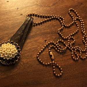 May include: A necklace with a pendant featuring a white flower with a brown metal frame. The necklace is made of a copper ball chain.