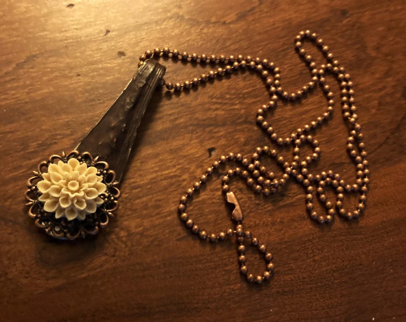 May include: A necklace with a pendant featuring a white flower with a brown metal frame. The necklace is made of a copper ball chain.