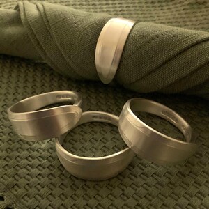 May include: Four silver napkin rings on a green textured surface. The rings are made of metal and have a smooth, polished finish.