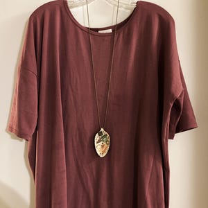 Op de afbeelding: Een loszittende, korte mouwen, maronkleurige t-shirt met een gouden kettingketting en een keramische hanger. De hanger heeft de vorm van een lepel en heeft een kleurrijk ontwerp.