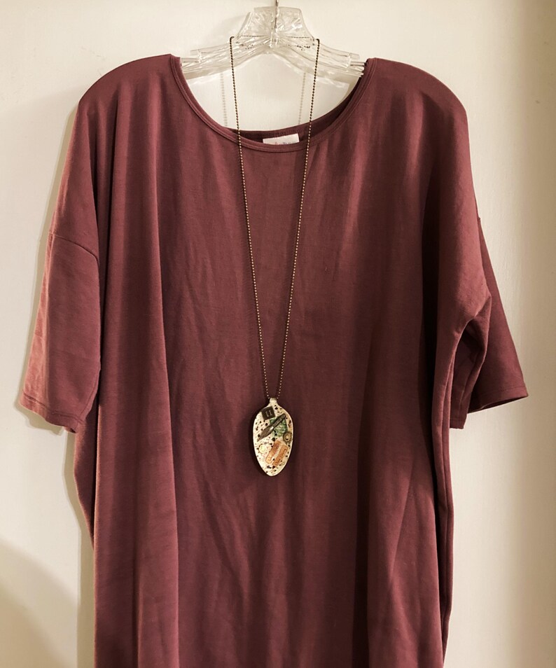 Op de afbeelding: Een loszittende, korte mouwen, maronkleurige t-shirt met een gouden kettingketting en een keramische hanger. De hanger heeft de vorm van een lepel en heeft een kleurrijk ontwerp.