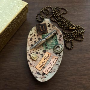 Op de afbeelding: Een keramische lepelvormige schaal versierd met een vintage pen, een gouden kroonknop en twee koperen platen met de tekst "Write Your Own Story". De schaal is ook versierd met een bruin leren patch met de letter "U" erop.