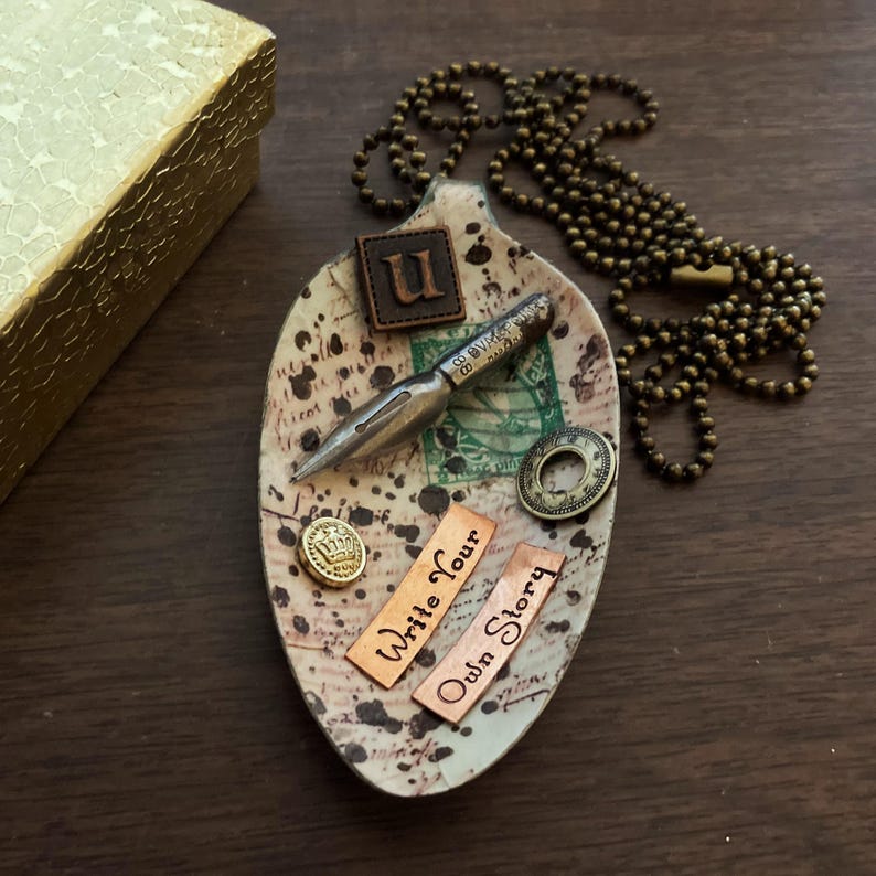 Op de afbeelding: Een keramische lepelvormige schaal versierd met een vintage pen, een gouden kroonknop en twee koperen platen met de tekst "Write Your Own Story". De schaal is ook versierd met een bruin leren patch met de letter "U" erop.