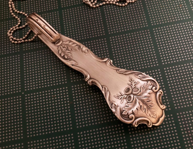 May include: A silver spoon necklace with an acorn design. The spoon is attached to a silver chain.