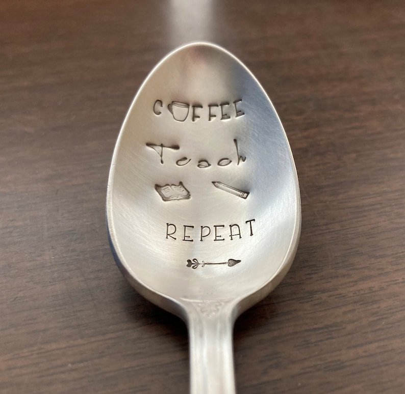 May include: A silver spoon with the words "Coffee Teach Repeat" engraved on it. There is a small coffee cup, a pencil, a book, and an arrow drawn on the spoon.