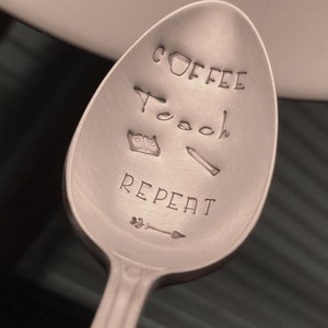 May include: A silver spoon with the words "Coffee Teach Repeat" engraved on it. There is a small coffee cup, a book, and a pencil drawn on the spoon.