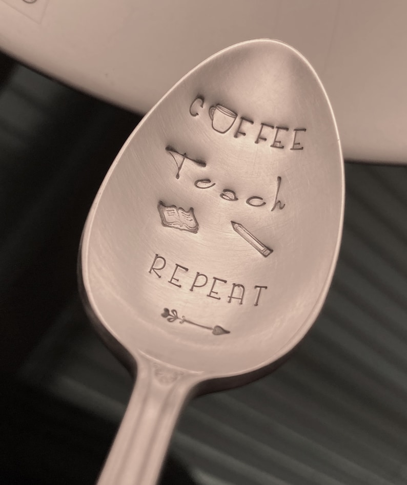 May include: A silver spoon with the words "Coffee Teach Repeat" engraved on it. There is a small coffee cup, a book, and a pencil drawn on the spoon.