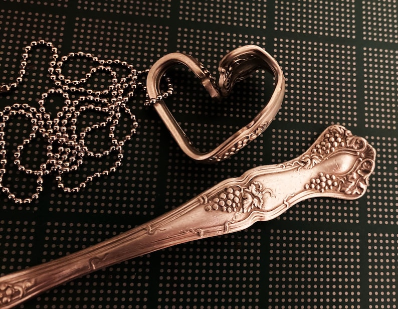 May include: A silver heart-shaped pendant necklace made from a bent spoon, with a silver chain and a silver spoon with a grapevine design.