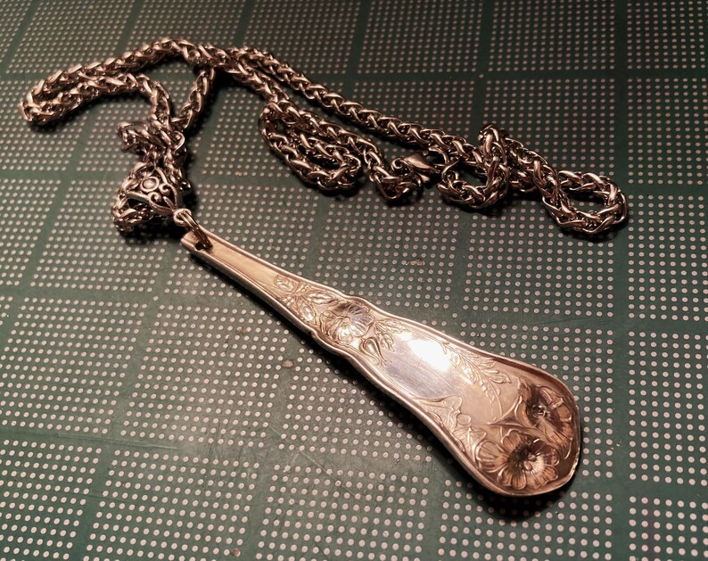 May include: A silver necklace with a detailed, ornate pendant made from a vintage spoon. The pendant has a floral design and is attached to a silver chain.