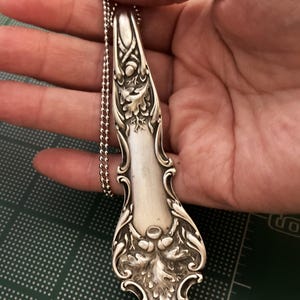 May include: A silver spoon pendant necklace with an ornate design of oak leaves and acorns. The spoon is attached to a silver ball chain.