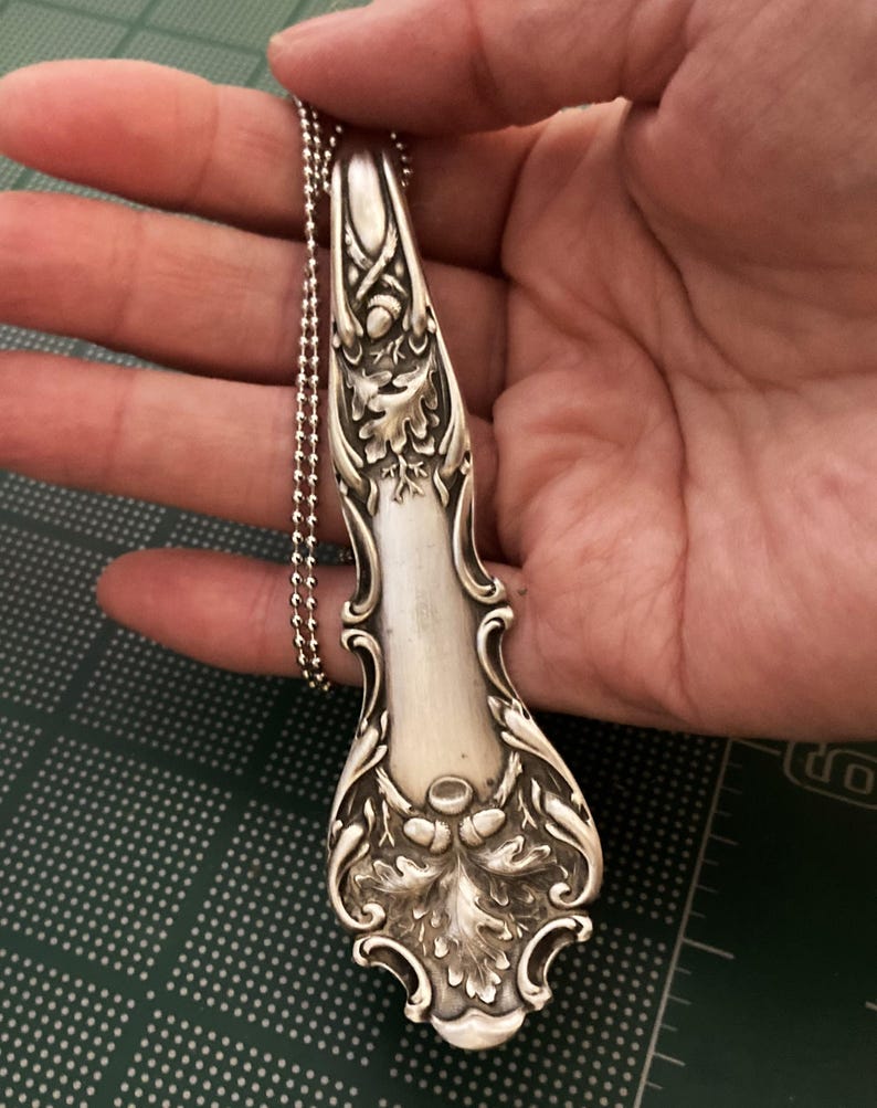 May include: A silver spoon pendant necklace with an ornate design of oak leaves and acorns. The spoon is attached to a silver ball chain.
